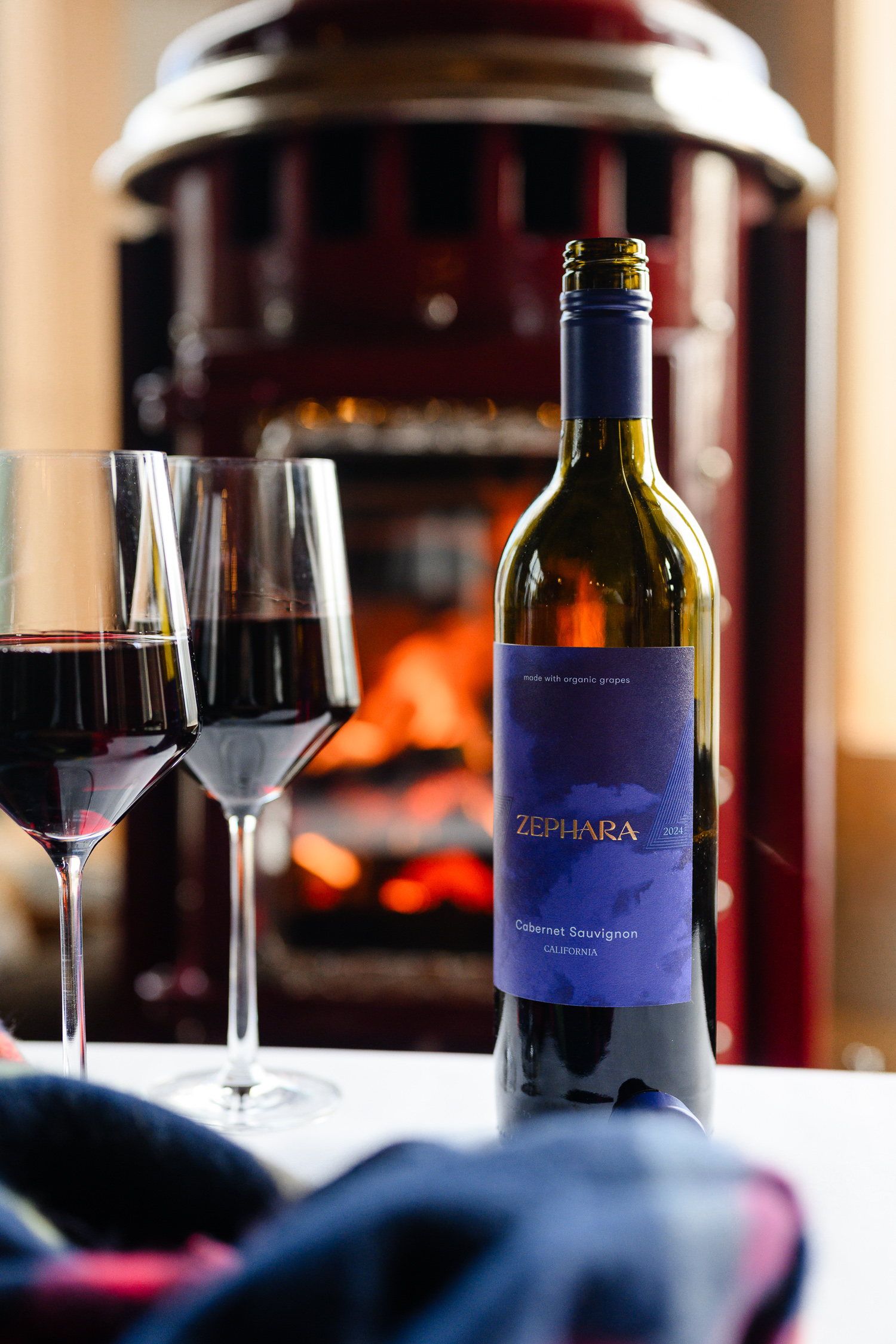 A bottle of Zephara Cabernet Sauvignon stands beside two glasses of red wine, with a glowing fireplace in the background. This cozy, intimate scene illustrates current branding trends that focus on lifestyle storytelling, captured by a brand photographer who elevates product warmth and connection