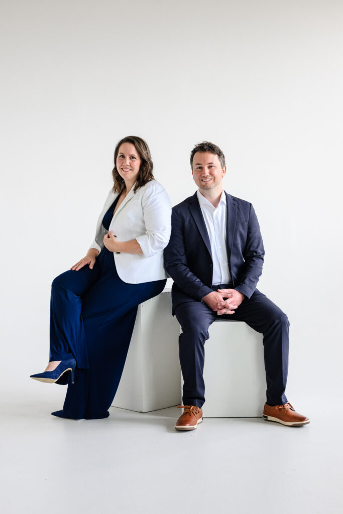 A professionally dressed man and woman sit on white studio cubes, smiling toward the camera in coordinated navy and white outfits as a branding photographer captures polished website photography during a denver branding session.