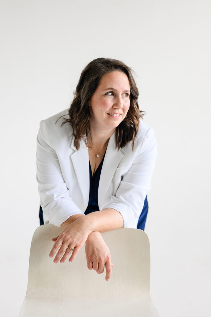 A woman in a white blazer leans over the back of a light wood chair against a seamless white studio backdrop as a branding photographer creates clean, minimalist website photography for a denver branding session.
