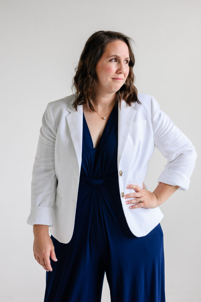 A woman in a white blazer layered over a navy jumpsuit stands with one hand on her hip, looking off-camera against a bright white studio backdrop in professional website photography by a branding photographer during a denver branding session.