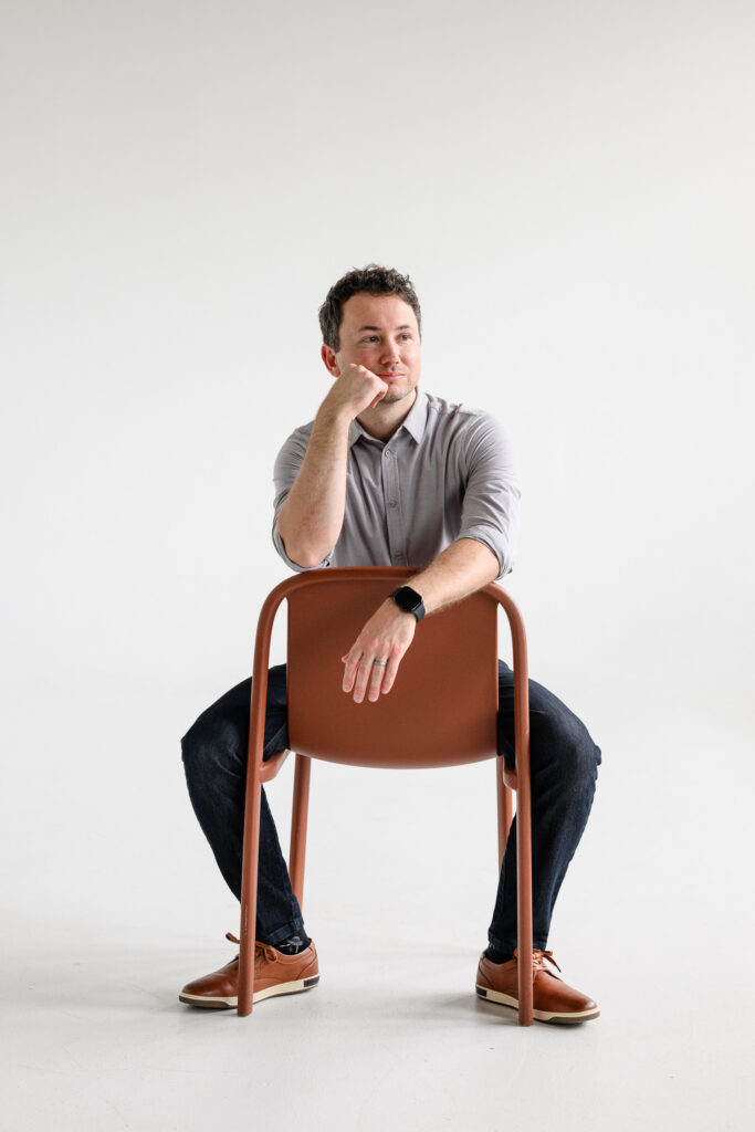 Resting his arm across the back of a brown chair, a casually dressed man looks thoughtfully off-camera against a seamless white studio backdrop in relaxed website photography by a branding photographer during a denver branding session.