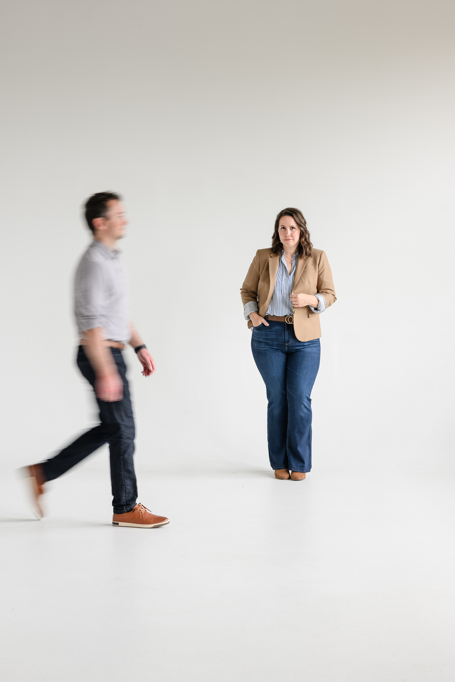 Intentional motion blur follows a man walking through the foreground while a woman in a tan blazer remains sharply in focus against a minimalist white studio backdrop, creating dynamic website photography captured by a branding photographer during a denver branding session.