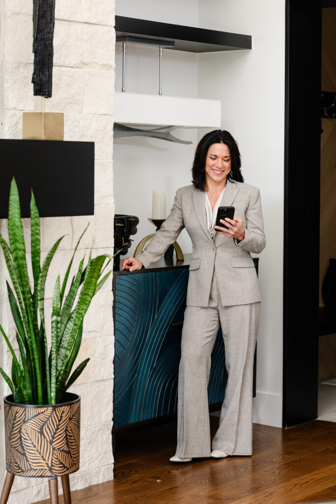 Professional woman in a tailored light gray pantsuit standing in a modern luxury home while checking her smartphone, surrounded by contemporary architectural details and stylish décor. This candid lifestyle portrait highlights confidence, professionalism, and everyday authenticity for brand photography, creating polished personal branding imagery captured by a Denver photographer.