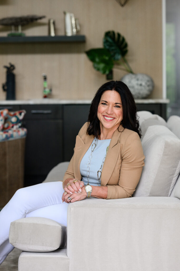 Professional woman in a tan blazer, light blue blouse, and white pants seated on a modern cream sofa in a contemporary office lounge, smiling warmly at the camera. This relaxed lifestyle portrait conveys confidence, professionalism, and approachability for brand photography, creating authentic personal branding imagery captured by a Denver photographer.