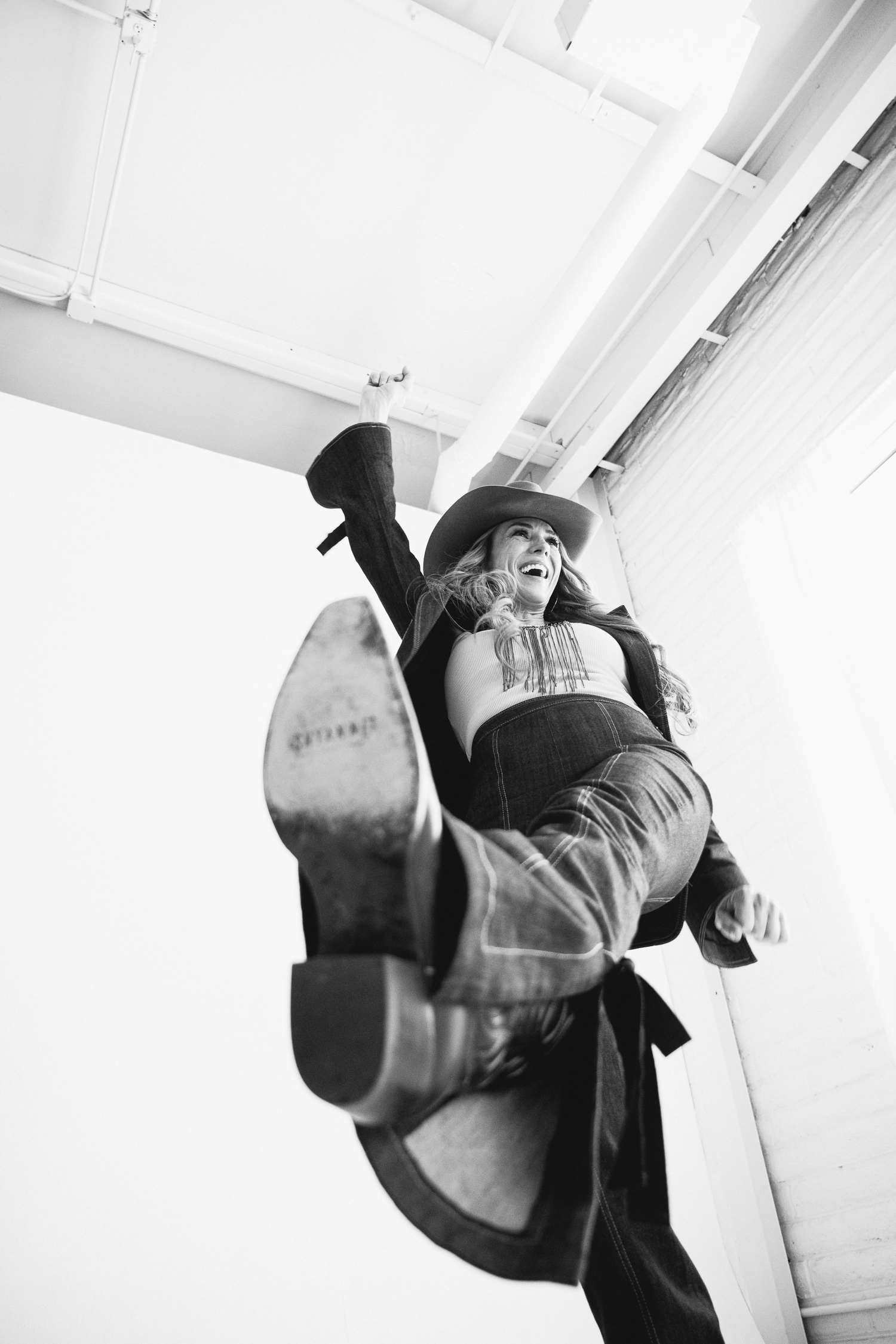 Black-and-white brandphotos of a confident woman in a cowboy hat and denim outfit striking a playful high-kick pose in a bright studio, captured by a denver branding photographer.