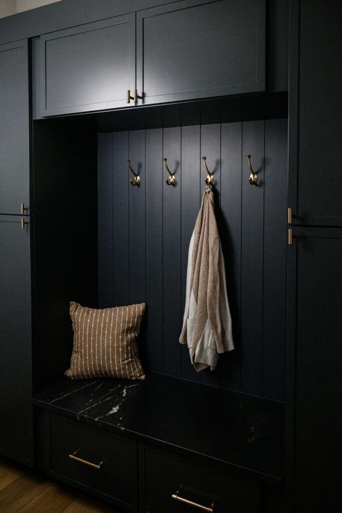 Moody built-in mudroom with charcoal cabinetry, brass hooks and hardware, marble bench, striped pillow, and hanging sweater captured through branding photography.