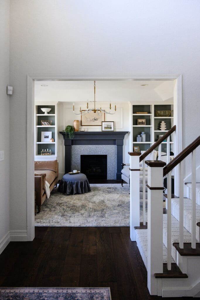 View from the entry into a layered living room with a dark fireplace, built-in shelving, chandelier, patterned rug, and staircase, captured for branding for interior designers.