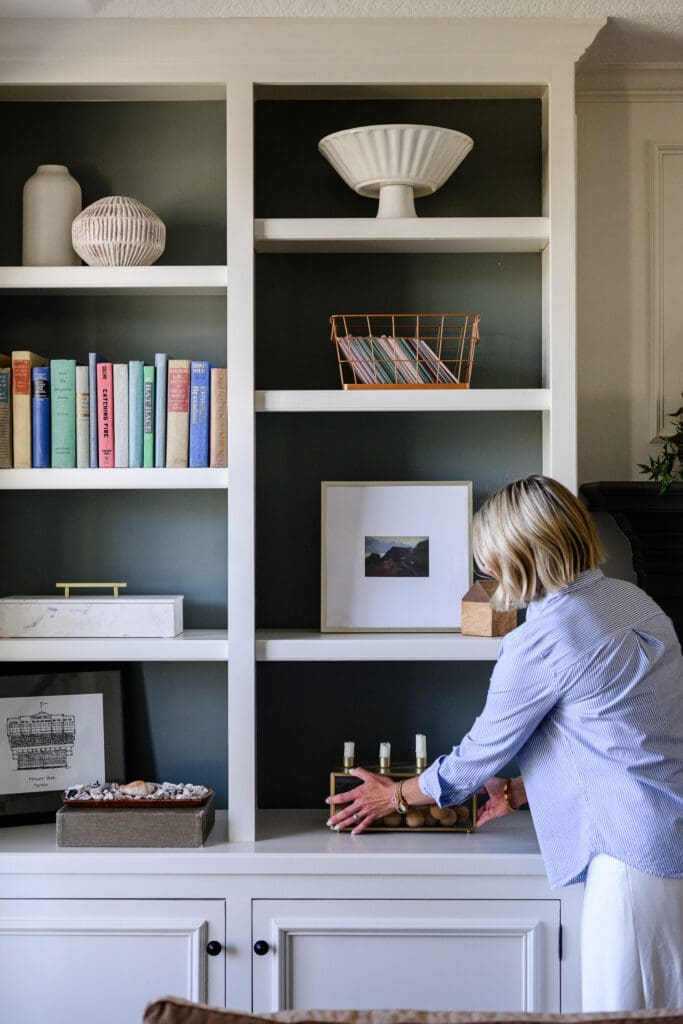 Interior designer arranging decor on dark-backed built-in shelves with books, ceramics, artwork, and collected objects during a branding photography session.